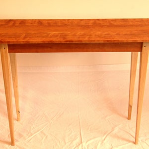 Puede incluir: Una mesa consola de madera con una tapa de color marrón claro y cuatro patas delgadas. La mesa está hecha de madera natural con un patrón de veta de madera clara.