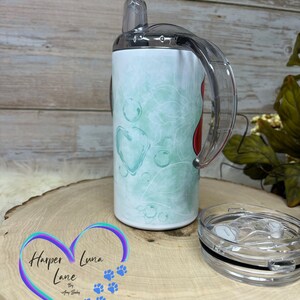 Puede incluir: Un vaso de acero inoxidable con un dise&ntilde;o en verde azulado y blanco y un coraz&oacute;n rojo. El vaso tiene una tapa transparente con una pajita y un asa transparente. El vaso est&aacute; sobre una superficie de madera con un logotipo que dice "Harper Luna Lane by Amy Selby".