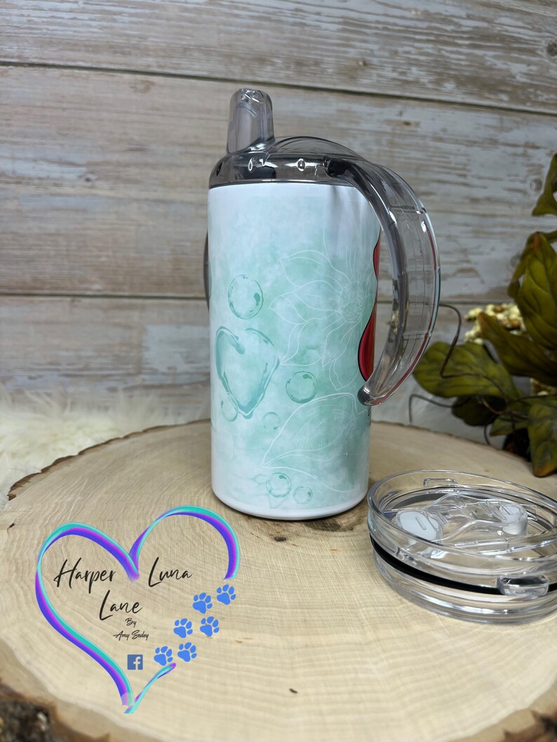 Puede incluir: Un vaso de acero inoxidable con un dise&ntilde;o en verde azulado y blanco y un coraz&oacute;n rojo. El vaso tiene una tapa transparente con una pajita y un asa transparente. El vaso est&aacute; sobre una superficie de madera con un logotipo que dice "Harper Luna Lane by Amy Selby".