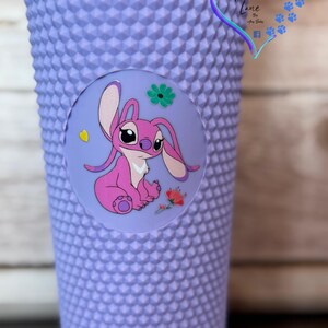 Puede incluir: Un vaso con tachuelas de color lavanda con una calcoman&iacute;a circular con un personaje de dibujos animados rosa, flores y un coraz&oacute;n amarillo. El vaso tiene una superficie texturizada y un logotipo en forma de coraz&oacute;n con el texto "Harper Luna Lane".