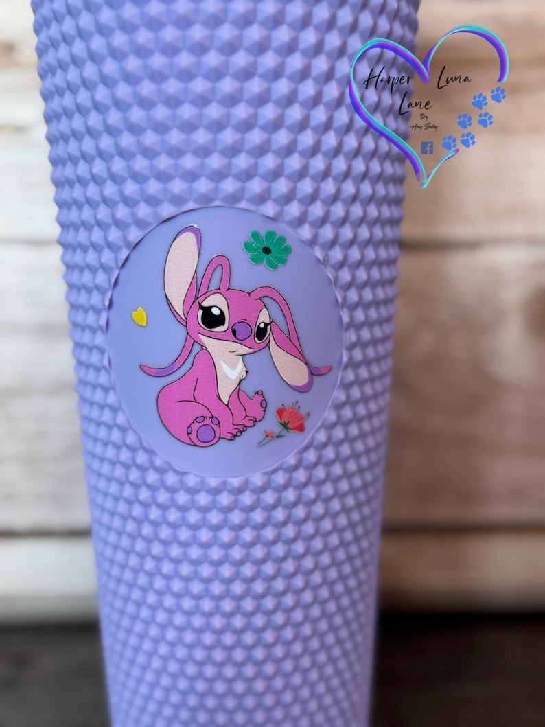 Puede incluir: Un vaso con tachuelas de color lavanda con una calcoman&iacute;a circular con un personaje de dibujos animados rosa, flores y un coraz&oacute;n amarillo. El vaso tiene una superficie texturizada y un logotipo en forma de coraz&oacute;n con el texto "Harper Luna Lane".