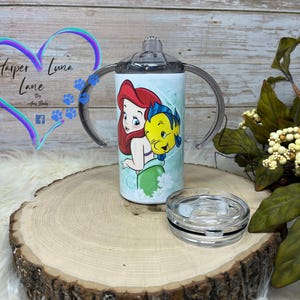 Puede incluir: Un vaso de acero inoxidable con una tapa transparente y dos asas. El vaso presenta una ilustraci&oacute;n colorida de Ariel de La Sirenita y Sebasti&aacute;n. El vaso est&aacute; sobre una superficie de madera.