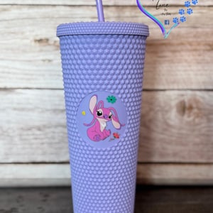 Puede incluir: Un vaso con tachuelas de color lavanda con tapa y pajita a juego. Una pegatina circular en la parte delantera presenta un personaje de dibujos animados rosa con una flor y una rosa. El vaso tiene el texto "Harper Luna Lane".