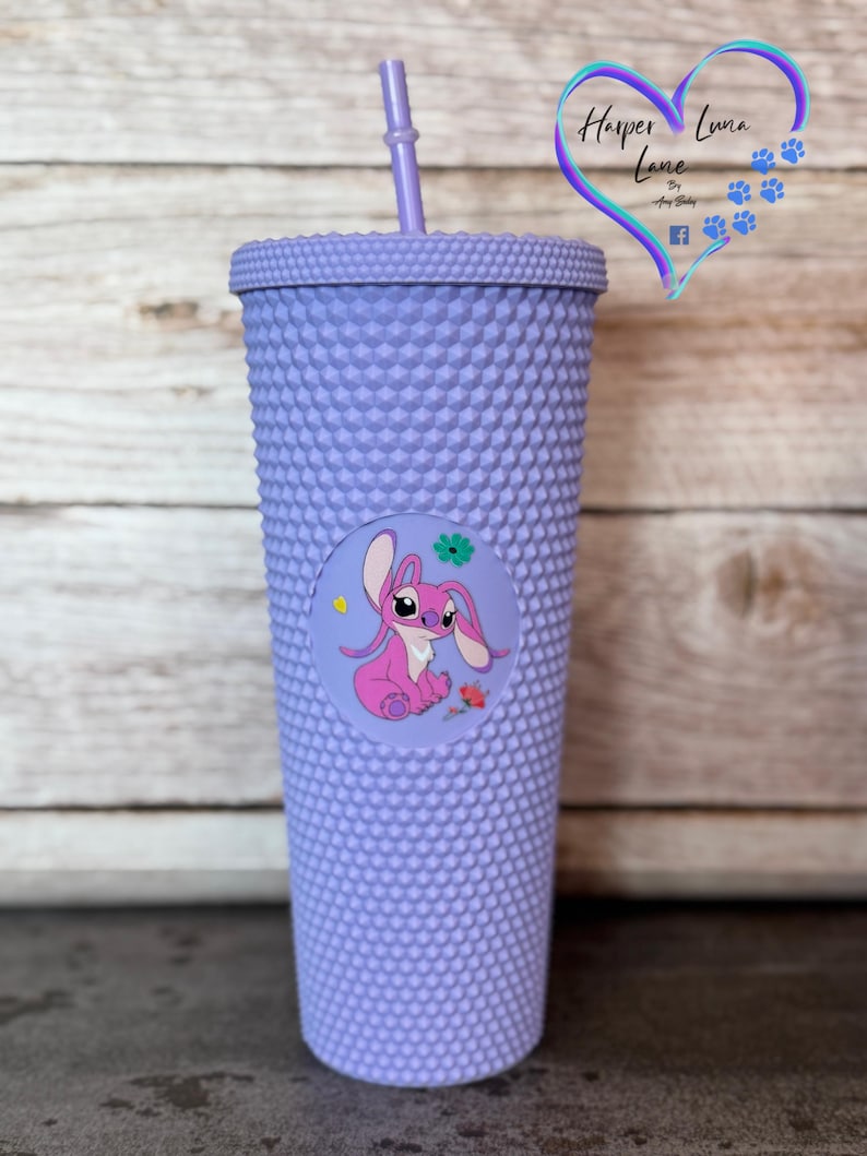 Puede incluir: Un vaso con tachuelas de color lavanda con tapa y pajita a juego. Una pegatina circular en la parte delantera presenta un personaje de dibujos animados rosa con una flor y una rosa. El vaso tiene el texto "Harper Luna Lane".