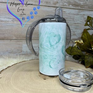 Puede incluir: Un vaso de acero inoxidable blanco con un dise&ntilde;o abstracto turquesa y blanco. El vaso tiene una tapa transparente con una pajita y dos asas. El vaso est&aacute; sobre una superficie de madera. El texto "Harper Luna Lane" es visible en el vaso.