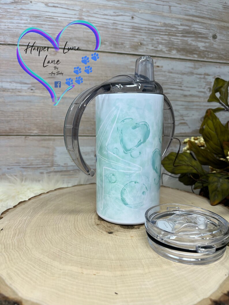 Puede incluir: Un vaso de acero inoxidable blanco con un dise&ntilde;o abstracto turquesa y blanco. El vaso tiene una tapa transparente con una pajita y dos asas. El vaso est&aacute; sobre una superficie de madera. El texto "Harper Luna Lane" es visible en el vaso.