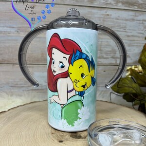 Puede incluir: Un vaso de acero inoxidable blanco con una tapa transparente y dos asas. El vaso presenta una imagen colorida de Ariel de La Sirenita y Sebasti&aacute;n. El vaso est&aacute; sobre una superficie de madera.