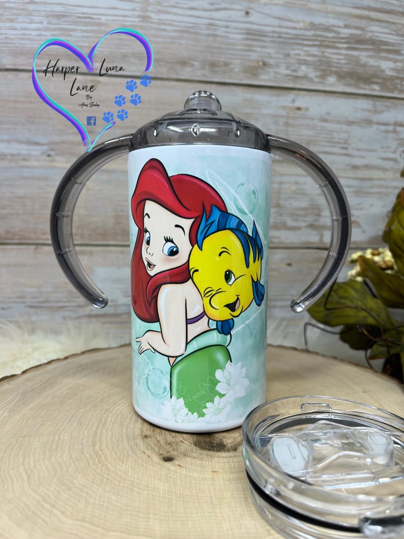 Puede incluir: Un vaso de acero inoxidable blanco con una tapa transparente y dos asas. El vaso presenta una imagen colorida de Ariel de La Sirenita y Sebasti&aacute;n. El vaso est&aacute; sobre una superficie de madera.