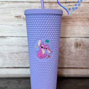 Puede incluir: Un vaso de color lavanda con tachuelas, con tapa y pajita a juego. Una calcoman&iacute;a circular presenta un personaje de dibujos animados rosa con orejas grandes y una flor. El vaso est&aacute; dise&ntilde;ado con una superficie texturizada.