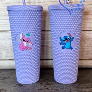 Puede incluir: Dos vasos morados con tachuelas y pajitas a juego. Cada vaso presenta un gr&aacute;fico circular de un personaje de dibujos animados. Uno muestra una criatura rosa con forma de conejo y el otro un personaje azul extraterrestre. Los vasos tienen una superficie texturizada.