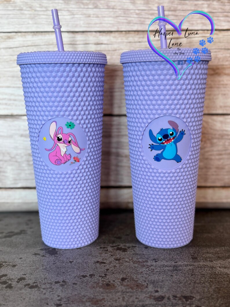 Puede incluir: Dos vasos morados con tachuelas y pajitas a juego. Cada vaso presenta un gr&aacute;fico circular de un personaje de dibujos animados. Uno muestra una criatura rosa con forma de conejo y el otro un personaje azul extraterrestre. Los vasos tienen una superficie texturizada.