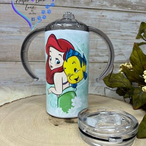 Puede incluir: Un vaso blanco con una tapa transparente y dos asas. El vaso presenta una ilustraci&oacute;n colorida de Ariel de La Sirenita y Sebasti&aacute;n. El vaso est&aacute; sobre una superficie de madera.