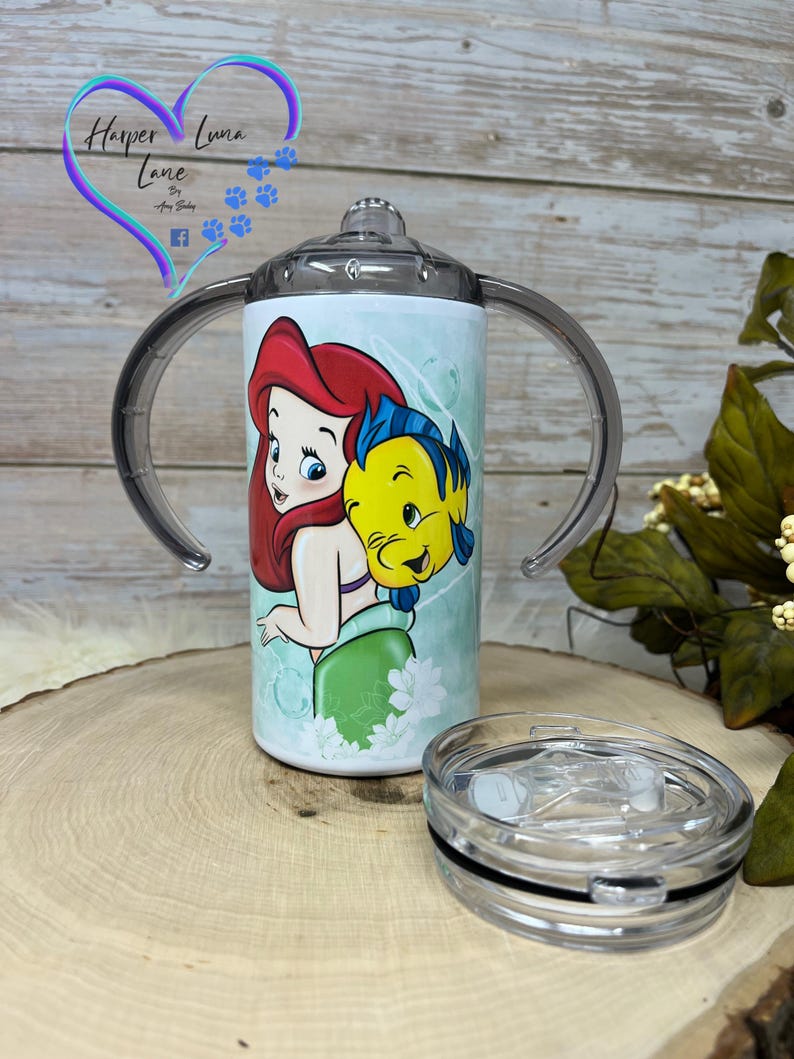 Puede incluir: Un vaso blanco con una tapa transparente y dos asas. El vaso presenta una ilustraci&oacute;n colorida de Ariel de La Sirenita y Sebasti&aacute;n. El vaso est&aacute; sobre una superficie de madera.