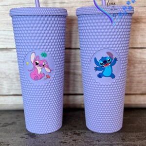 Puede incluir: Dos vasos de color lavanda con tachuelas y pajitas a juego. Cada vaso presenta un gr&aacute;fico circular de un personaje de dibujos animados. Uno tiene un personaje rosa con forma de conejo, el otro un personaje azul con forma de extraterrestre.
