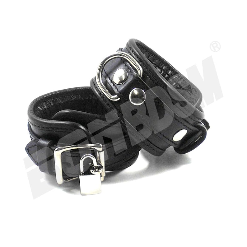 May include: A pair of black leather wrist cuffs with a silver lock and metal hardware. The cuffs are connected by a silver chain.