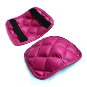 May include: Two pink quilted leather elbow pads with black elastic straps. The pads are designed to provide comfort and support for the elbows.