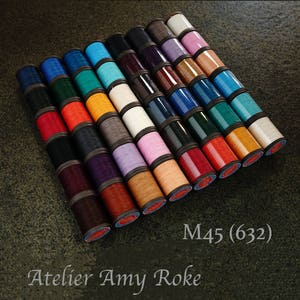 Puede incluir: Una colección de 24 carretes de hilo en varios colores, incluyendo rojo, naranja, amarillo, verde, azul, morado, rosa y blanco. Los carretes están dispuestos en forma de abanico y están etiquetados con "M45 (632)" y "Atelier Amy Roko".