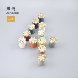 Può includere: Una collezione di 12 bobine di filo di vari colori, tra cui beige, marrone, verde e blu. Ogni bobina è etichettata con il numero "432" e il testo "Au Chinois".