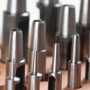 May include: A set of metal hole punches of various sizes, arranged in a row. The punches are silver in color and have a sharp, pointed tip. They are used for creating holes in leather, paper, or other materials.