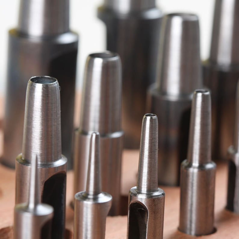 May include: A set of metal hole punches of various sizes, arranged in a row. The punches are silver in color and have a sharp, pointed tip. They are used for creating holes in leather, paper, or other materials.