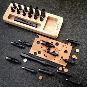 May include: A set of leather hole punches in various sizes, with a wooden storage tray labeled "Art & Soul". The punches are arranged in the tray and some are scattered on a brown surface with a grid of holes.