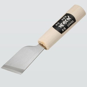 Pode incluir: Uma pequena faca japonesa de trabalho de couro, de cor prateada, com cabo de madeira. O cabo tem texto japonês que diz "真鍮製", que se traduz como "feito de latão".