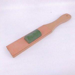 以下が含まれることがあります： 薄いピンクの革の表面と緑色の砥石が付いた木製のストロップ。ストロップには木製のハンドルが付いています。
