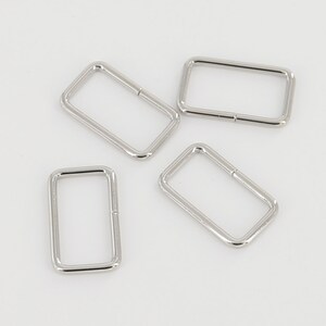Peut inclure: Quatre anneaux en D en métal argenté, également appelés anneaux de saut, sont disposés sur une surface blanche. Les anneaux sont de forme rectangulaire et ont une finition lisse et polie.