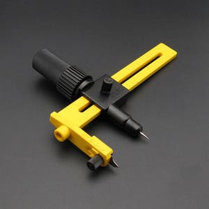 Pode incluir: Um cortador circular amarelo e preto com uma pega preta e um dial rotativo. O cortador é projetado para cortar círculos em materiais como papel, tecido e couro.