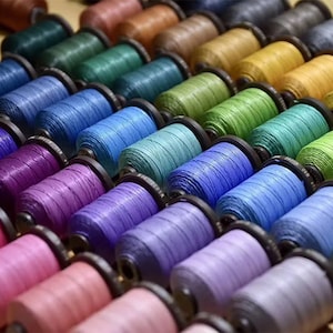 Könnte beinhalten: Nahaufnahme von Spulen mit farbigem Garn, die in Reihen angeordnet sind. Das Garn ist um Spulen mit Metallkappen gewickelt. Die Farben des Garns umfassen Lila, Blau, Grün, Braun und Rosa.