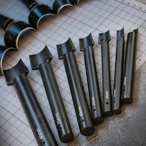 Puede incluir: Conjunto de ocho herramientas de punzonado de cuero negras de diferentes tamaños, que van desde 15 mm hasta 40 mm. Cada herramienta tiene un borde curvo y un mango.