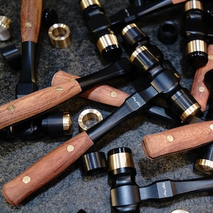 以下が含まれることがあります： 木製のハンドルが付いた黒と金のハンマー型のツール6個セット。ツールは滑らかで磨かれた仕上げで、暗い表面に配置されています。