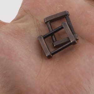 K&ouml;nnte beinhalten: Zwei Metallschnallen in dunkelbrauner Farbe. Die Schnallen sind rechteckig mit einer quadratischen &Ouml;ffnung in der Mitte. Die Schnallen sind &uuml;bereinander gestapelt.