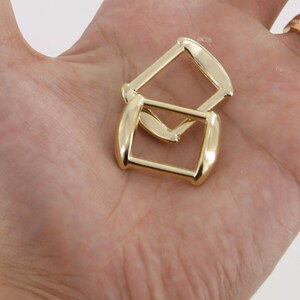 May include: Two gold-colored metal belt buckle loops. The loops are rectangular with rounded edges.
