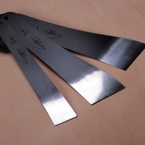 Puede incluir: Cuatro cuchillas de metal plateado con acabado cepillado y una firma negra en cada cuchilla. Las cuchillas están dispuestas en forma de abanico.