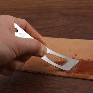 Könnte beinhalten: Ein silbernes Metallwerkzeug mit einem Loch im Griff und einer gebogenen Klinge wird verwendet, um eine braune Substanz auf einem Stück Leder zu verteilen.