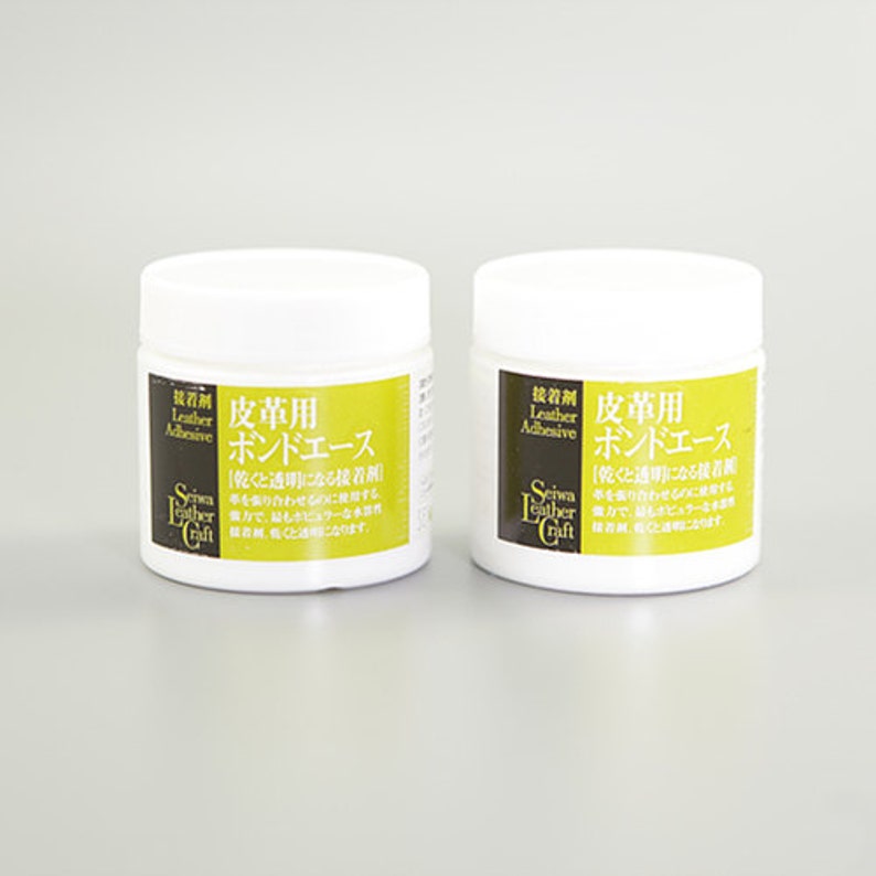 K&ouml;nnte beinhalten: Zwei wei&szlig;e Gl&auml;ser mit Lederkleber, beschriftet mit "Leather Adhesive" und "皮革用  ボンドエース" in Japanisch. Die Gl&auml;ser stehen auf einer wei&szlig;en Oberfl&auml;che.