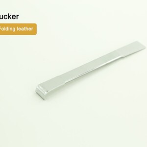 Può includere: Uno strumento in metallo argentato con un'estremità arrotondata, etichettato "Folding leather" e "Tucker".