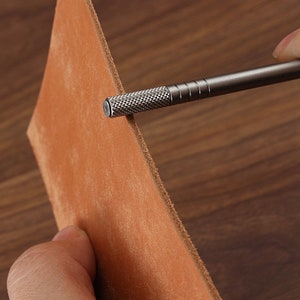May include: A close-up of a metal leather tool being used to punch a hole in a piece of brown leather.