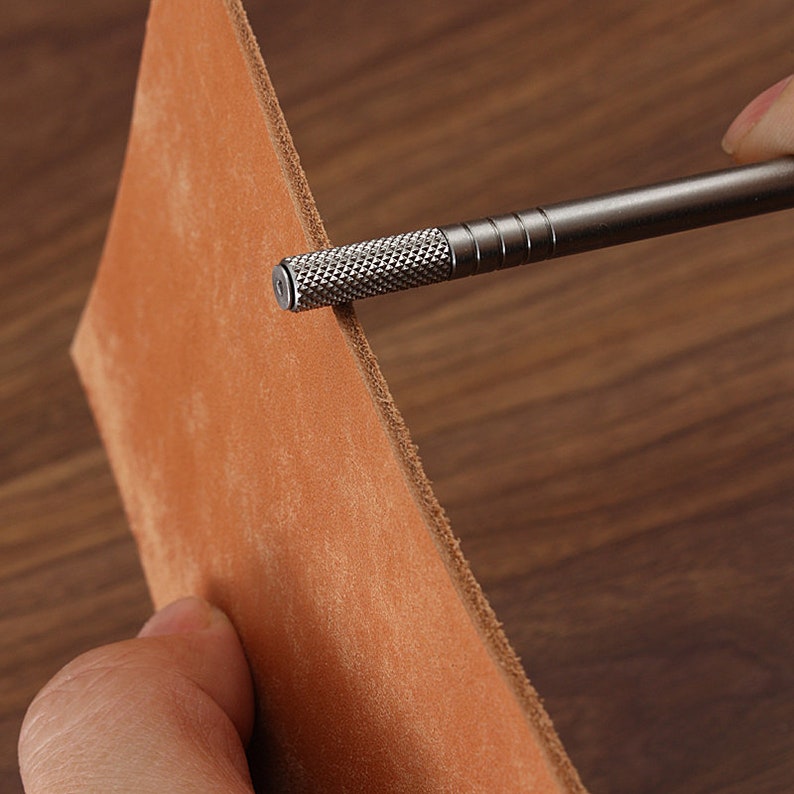 May include: A close-up of a metal leather tool being used to punch a hole in a piece of brown leather.