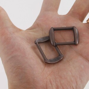Puede incluir: Dos hebillas rectangulares de color bronce antiguo con bordes redondeados. Las hebillas son de metal y se muestran en una mano. Las hebillas son probablemente para usar en marroquiner&iacute;a u otros proyectos de artesan&iacute;a.