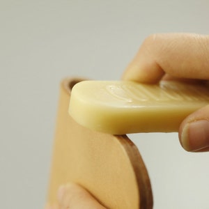 May include: A hand applying a bar of light brown leather conditioner to a piece of light brown leather.