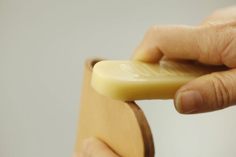 May include: A hand applying a bar of light brown leather conditioner to a piece of light brown leather.