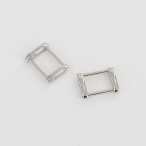 Puede incluir: Dos hebillas deslizantes de metal plateado. Las hebillas son de forma rectangular con esquinas redondeadas.