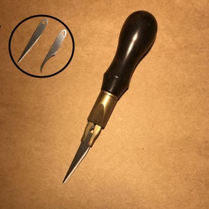 Puede incluir: Una herramienta de artesanía de cuero con mango negro, cuerpo de latón y hoja intercambiable. Se muestran dos hojas adicionales en la imagen.