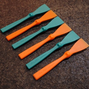 Könnte beinhalten: Sechs orangefarbene und blaugrüne Kunststoff-Kleberabzieher mit dem Aufdruck "Amy Robs" und "Glue Paddle, Italy".