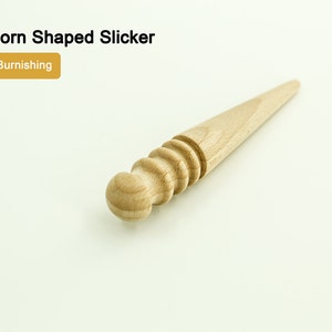 Op de afbeelding: Een houten maisvormige slicker-tool voor het gladstrijken van leer. De tool heeft een afgerond uiteinde en een getextureerd, geribbeld lichaam.