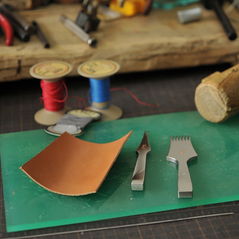 Peut inclure: Un tapis de coupe vert avec deux outils de travail du cuir, un poin&ccedil;on &agrave; cuir et un ciseau &agrave; piquer, et un morceau de cuir brun.