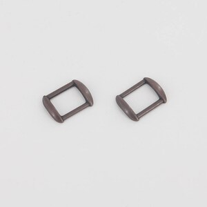 Puede incluir: Dos hebillas rectangulares de color bronce con bordes redondeados se muestran sobre un fondo blanco. Las hebillas son id&eacute;nticas en forma y tama&ntilde;o, lo que sugiere que est&aacute;n dise&ntilde;adas para usarse en pares, posiblemente para un cintur&oacute;n o una correa.