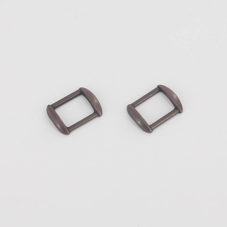 Puede incluir: Dos hebillas rectangulares de color bronce con bordes redondeados se muestran sobre un fondo blanco. Las hebillas son id&eacute;nticas en forma y tama&ntilde;o, lo que sugiere que est&aacute;n dise&ntilde;adas para usarse en pares, posiblemente para un cintur&oacute;n o una correa.